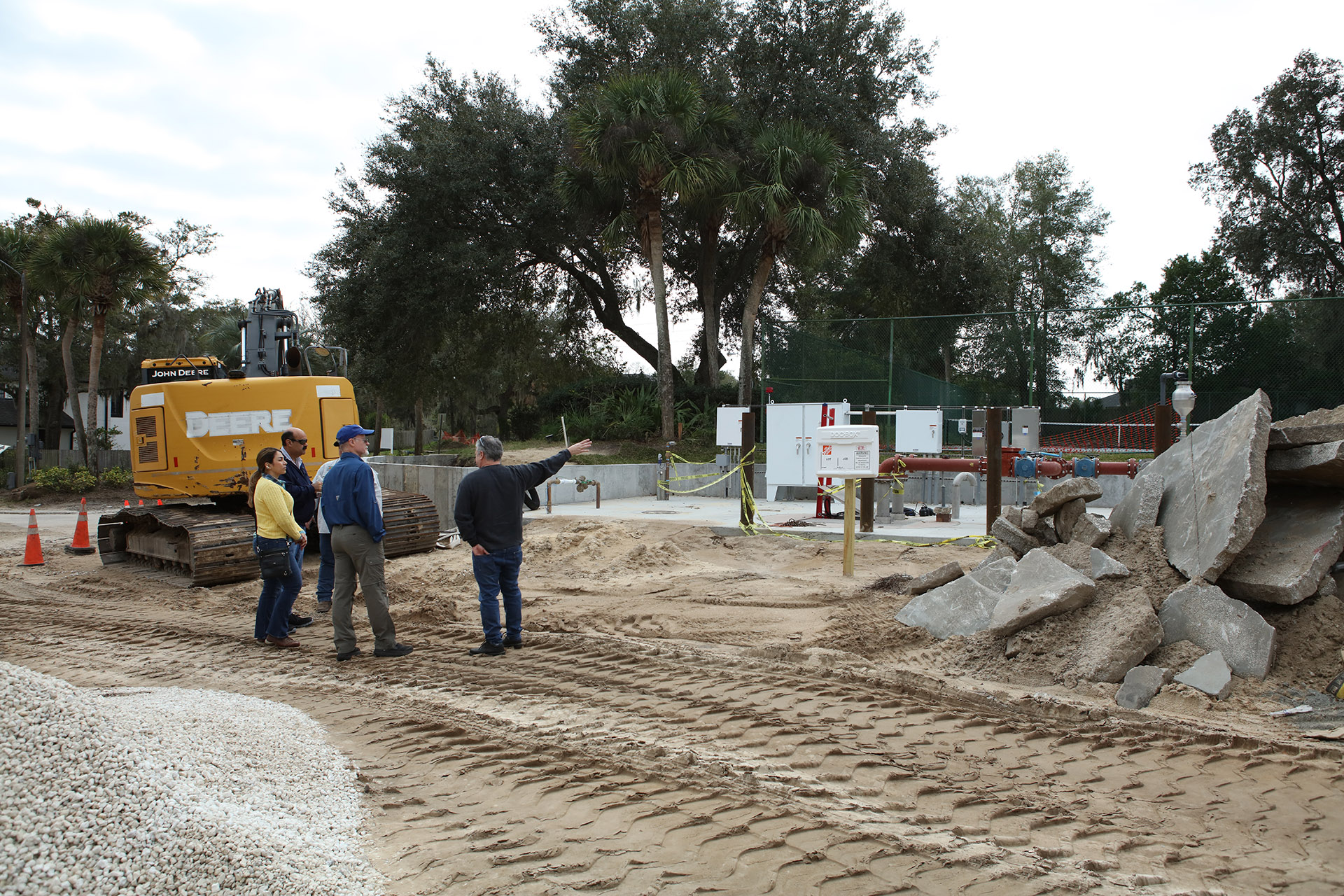 Four men and one woman standing in a dirt path created by an excavator. One main points at system of pipes a few yards ahead.